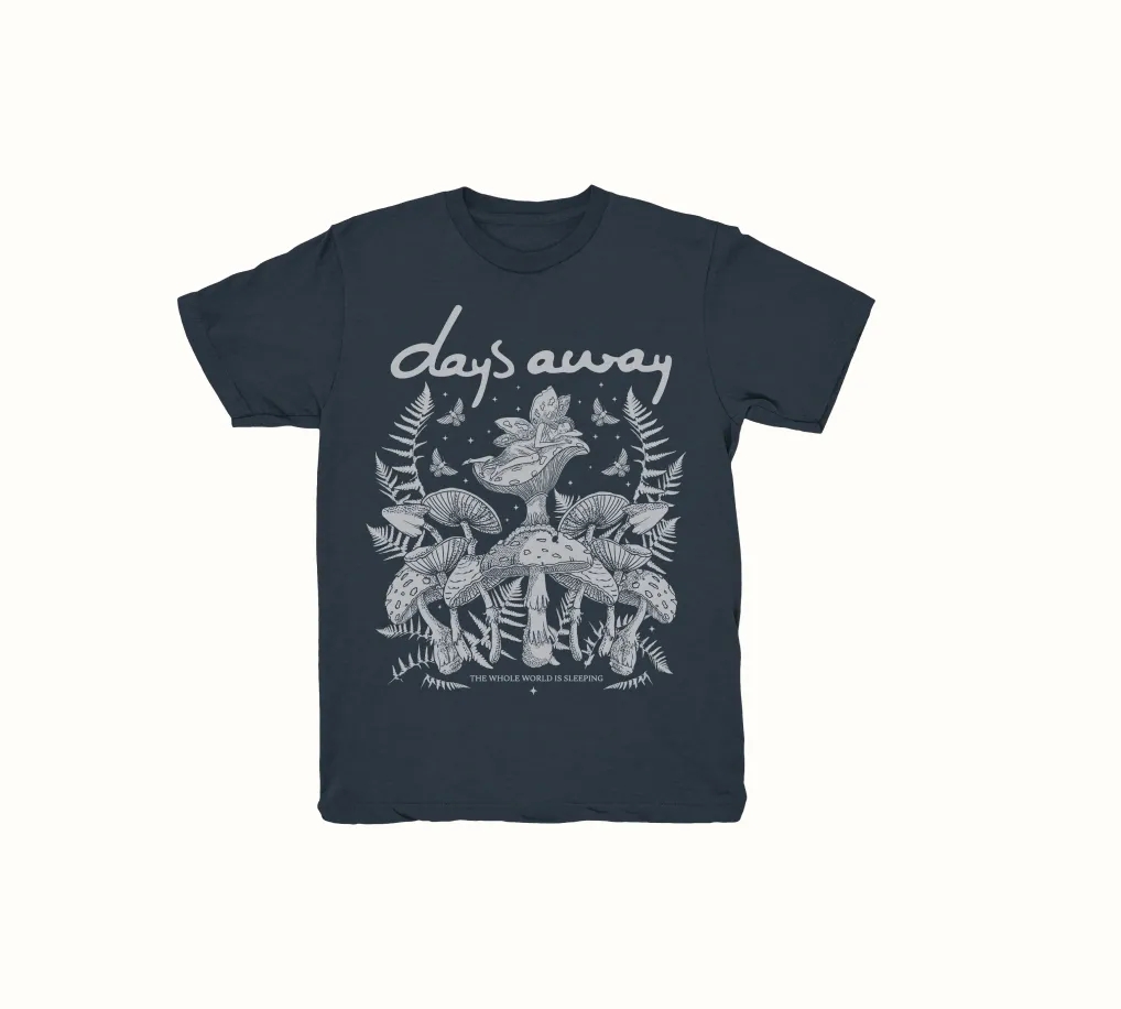 Front of a t-shirt featuring the Days Away logo at the chest, with an illustration of a fairy resting on a mushroom, surrounded by other mushrooms and ferns, creating a whimsical and nature-inspired design.